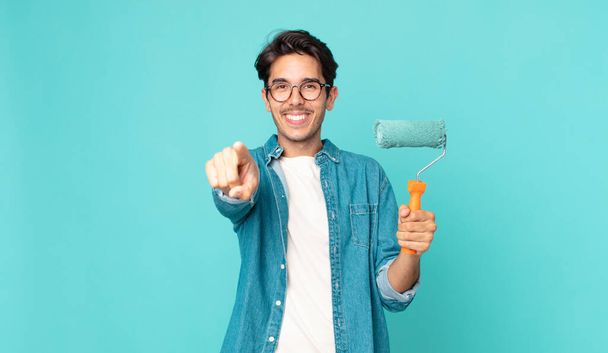 young hispanic man pointing at camera choosing you and holding a paint roller - Foto, Bild