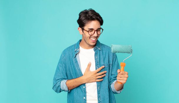 young hispanic man laughing out loud at some hilarious joke and holding a paint roller - Fotoğraf, Görsel