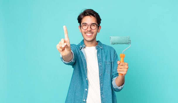 young hispanic man smiling proudly and confidently making number one and holding a paint roller - Foto, afbeelding