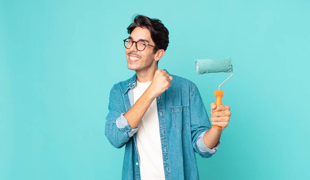 young hispanic man feeling happy and facing a challenge or celebrating and holding a paint roller - Photo, image