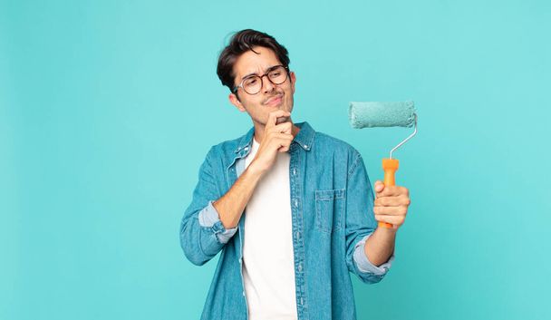 young hispanic man thinking, feeling doubtful and confused and holding a paint roller - 写真・画像