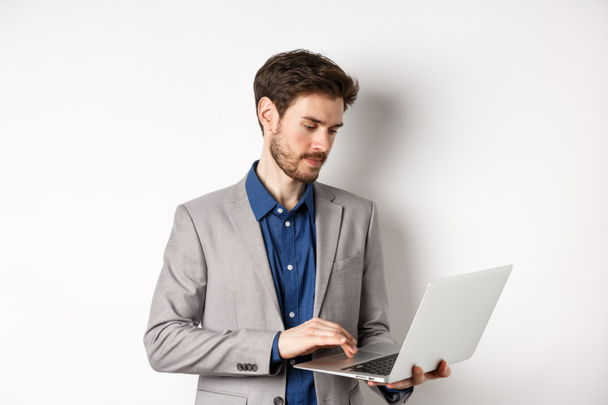Schöner männlicher Unternehmer, der am Laptop arbeitet, ernst auf den Bildschirm schaut und vor weißem Hintergrund steht - Foto, Bild