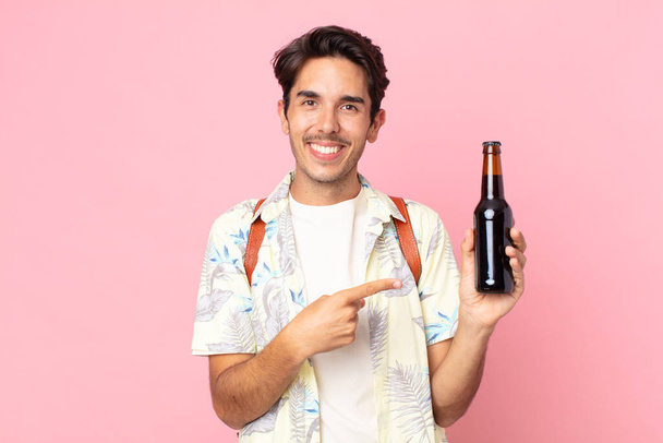 young hispanic man smiling cheerfully, feeling happy and pointing to the side and holding a bottle of beer - Foto, Imagen