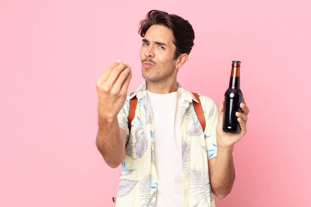 young hispanic man making capice or money gesture, telling you to pay and holding a bottle of beer - Foto, Imagen