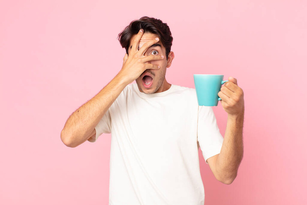 young hispanic man looking shocked, scared or terrified, covering face with hand and holding a coffee mug - Zdjęcie, obraz