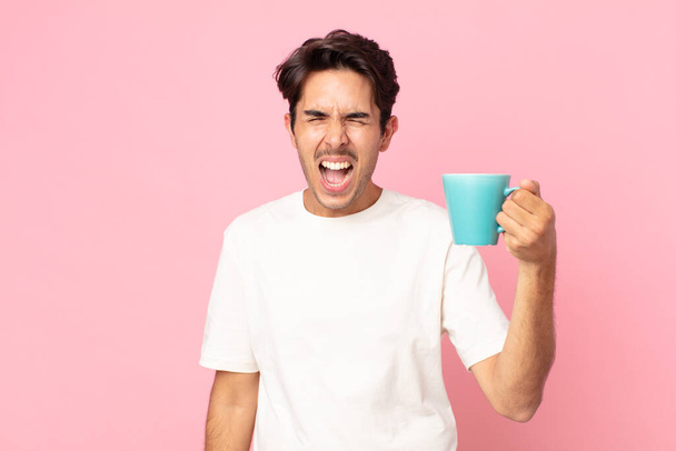young hispanic man shouting aggressively, looking very angry and holding a coffee mug - Foto, imagen