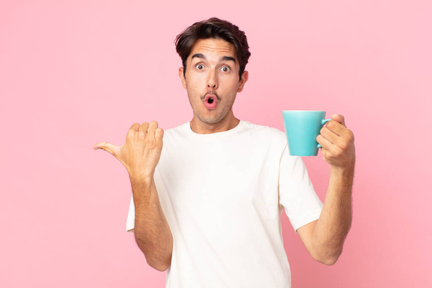 young hispanic man looking astonished in disbelief and holding a coffee mug - Zdjęcie, obraz