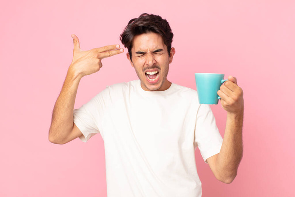 young hispanic man looking unhappy and stressed, suicide gesture making gun sign and holding a coffee mug - Photo, image