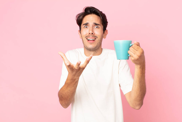 young hispanic man looking desperate, frustrated and stressed and holding a coffee mug - Фото, изображение