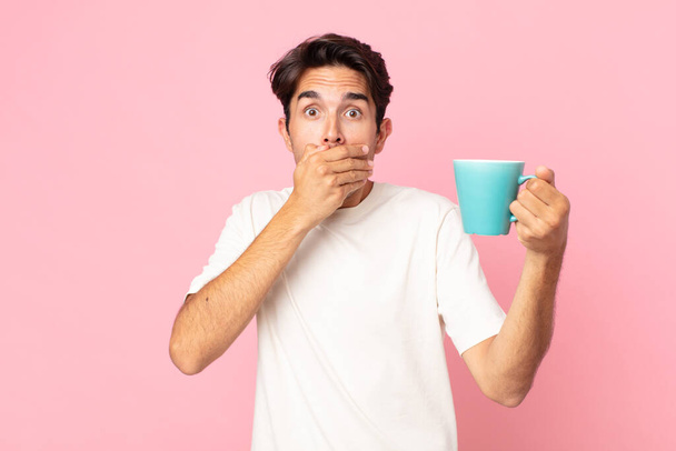 young hispanic man covering mouth with hands with a shocked and holding a coffee mug - Foto, imagen