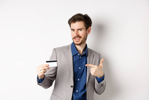 Επιτυχημένος επιχειρηματίας που δείχνει σε πλαστική πιστωτική κάρτα και χαμογελώντας, συνιστώντας τράπεζα, στέκεται σε λευκό φόντο - Φωτογραφία, εικόνα
