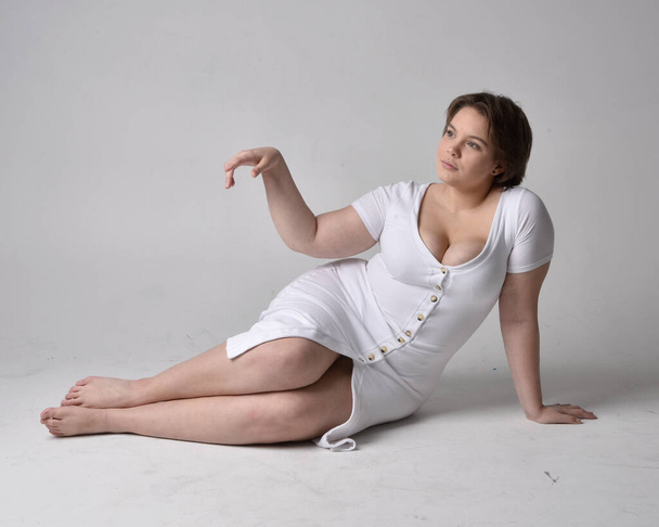 Full length portrait of young plus sized woman with short brunette hair,  wearing l tight white body con dress,  kneeling pose with gestural hands on ground with light studio background. - Fotografie, Obrázek