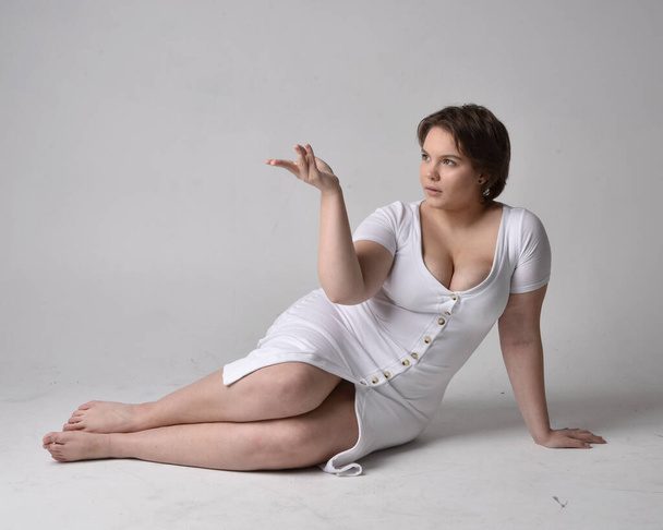 Full length portrait of young plus sized woman with short brunette hair,  wearing l tight white body con dress,  kneeling pose with gestural hands on ground with light studio background. - Photo, Image