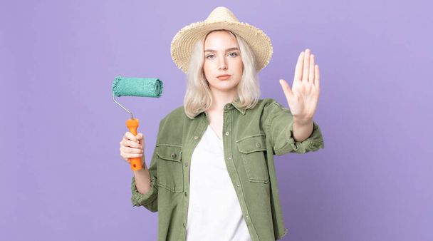 young pretty albino woman looking serious showing open palm making stop gesture and holding a roller paint - Zdjęcie, obraz