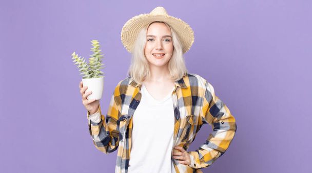 young pretty albino woman smiling happily with a hand on hip and confident and holding a houseplant cactus - Foto, Imagem