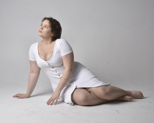 Full length portrait of young plus sized woman with short brunette hair,  wearing l tight white body con dress,  kneeling pose with gestural hands on ground with light studio background. - Zdjęcie, obraz