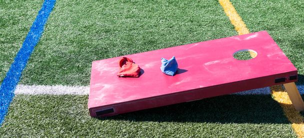 Een rode zelfgemaakte maïsgat boon tas gooien spel op een groene grasveld met rode en blauwe zakken op de top. - Foto, afbeelding
