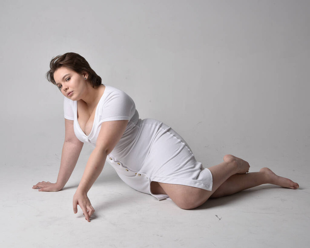 Full length portrait of young plus sized woman with short brunette hair,  wearing l tight white body con dress,  kneeling pose with gestural hands on ground with light studio background. - 写真・画像