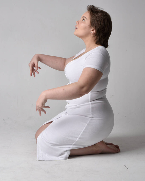 Full length portrait of young plus sized woman with short brunette hair,  wearing l tight white body con dress,  kneeling pose with gestural hands on ground with light studio background. - Fotó, kép