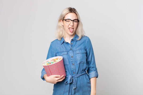 young pretty albino woman with cheerful and rebellious attitude, joking and sticking tongue out with a pop corns bucket - Photo, Image