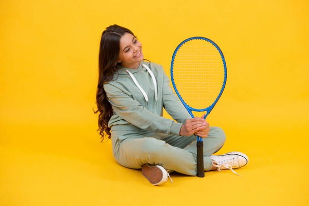 Spor kıyafetleri içinde neşeli bir genç kız sarı arka planda tenis raketi tutuyor. - Fotoğraf, Görsel