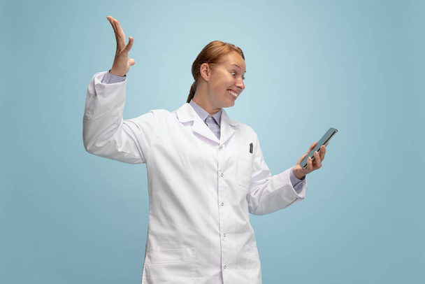 Portrait of female doctor, therapeutic or medical advisor with smartphone screen isolated on blue background. Funny happy emotions. Concept of healthcare, care medicine and humor - Photo, image