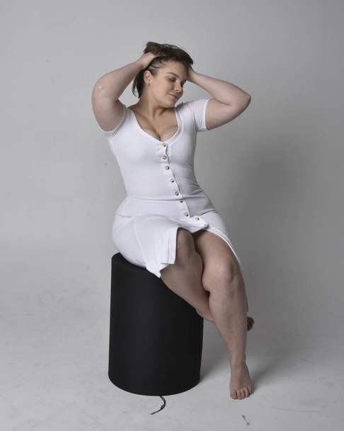 Full length portrait of young plus sized woman with short brunette hair,  wearing tight white body con dress,   sitting pose on chair with gestural hands with light studio background. - Фото, зображення