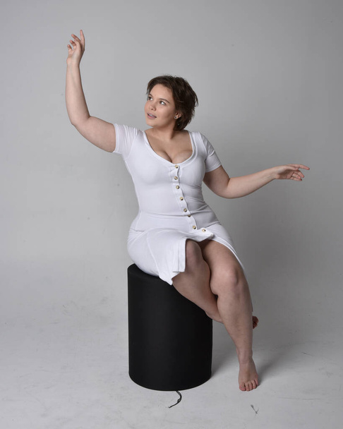 Ganzlanges Porträt einer jungen Frau mit kurzen brünetten Haaren, die ein enges weißes Body-Con-Kleid trägt und auf einem Stuhl mit gestikulierenden Händen vor hellem Studiohintergrund posiert. - Foto, Bild