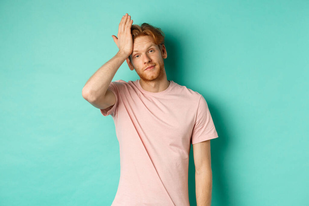 Annoyed and bothered redhead male model showing facepalm gesture, standing over mint background - Photo, Image