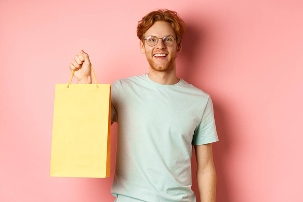 Hezký mladý muž kupující dárky, držící nákupní tašku a usmívající se na kameru, stojící nad růžovým pozadím - Fotografie, Obrázek