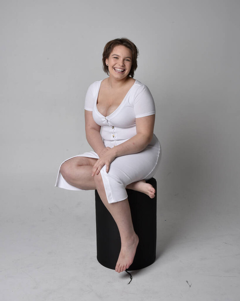 Full length portrait of young plus sized woman with short brunette hair,  wearing tight white body con dress,   sitting pose on chair with gestural hands with light studio background. - Fotografie, Obrázek