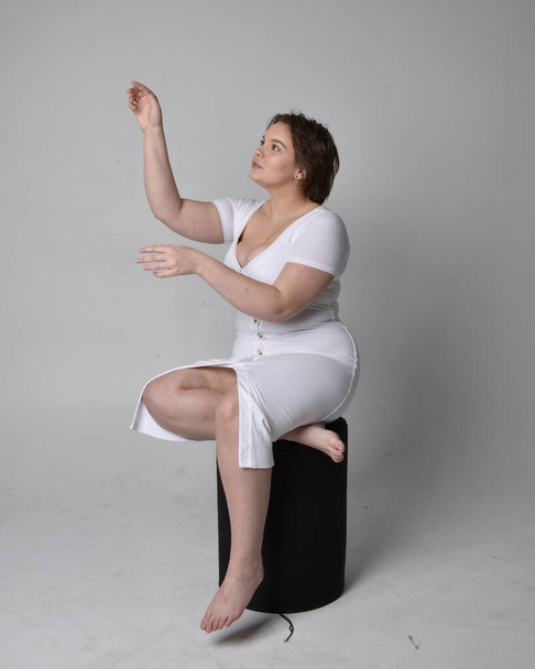 Полноразмерный портрет молодой женщины с короткими волосами брюнетки, в обтягивающем белом платье для обмана тела, сидящей на стуле с жестикулирующими руками на светлом студийном фоне. - Фото, изображение