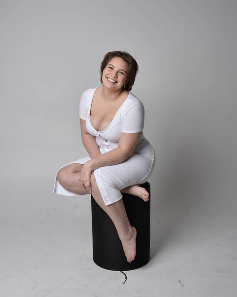 Ganzlanges Porträt einer jungen Frau mit kurzen brünetten Haaren, die ein enges weißes Body-Con-Kleid trägt und auf einem Stuhl mit gestikulierenden Händen vor hellem Studiohintergrund posiert. - Foto, Bild