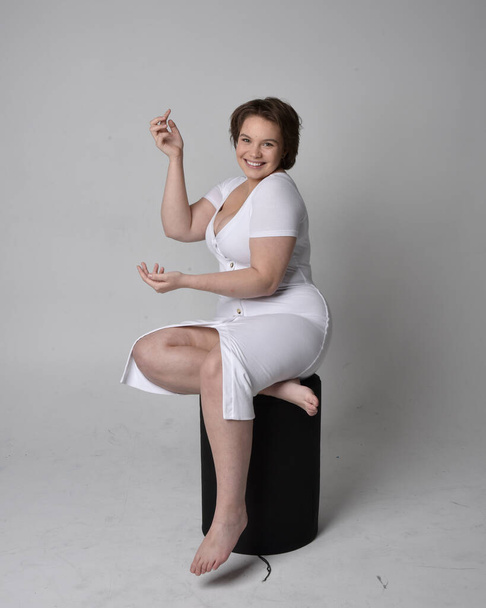 Full length portrait of young plus size woman with short brunette hair, wearing tight white body con dress, sitting pose on chair with gebural hands with light studio background. - Foto, afbeelding