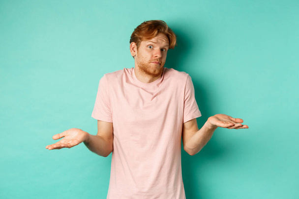 Young redhead guy in t-shirt know nothing, shrugging shoudlers with hands spread sideways, standing confused over turquoise background - Photo, Image