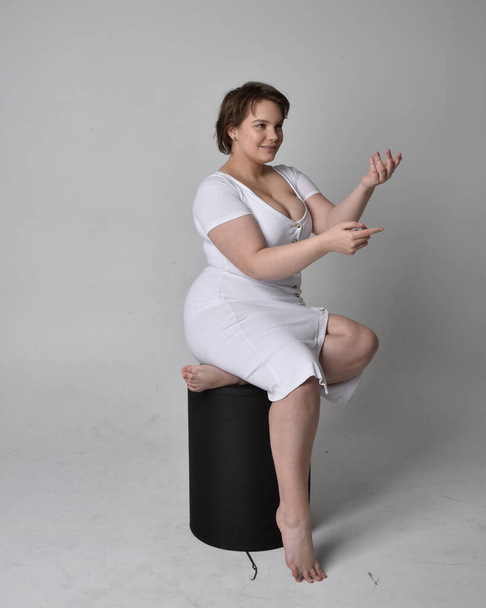 Full length portrait of young plus sized woman with short brunette hair,  wearing tight white body con dress,   sitting pose on chair with gestural hands with light studio background. - Photo, Image