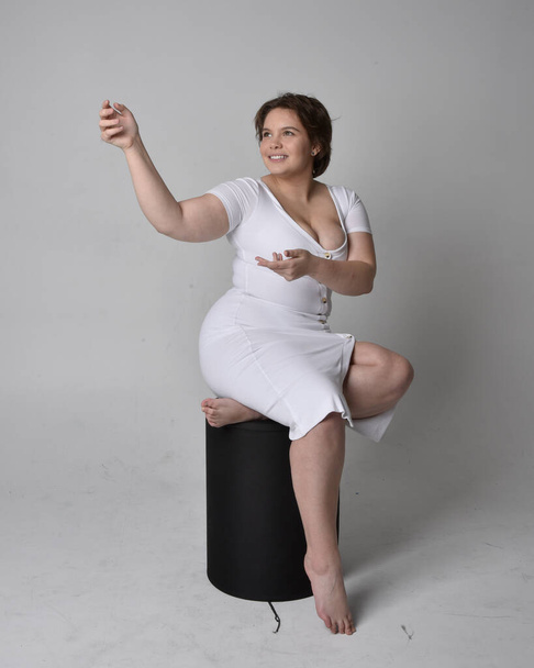 Full length portrait of young plus size woman with short brunette hair, wearing tight white body con dress, sitting pose on chair with gebural hands with light studio background. - Foto, afbeelding