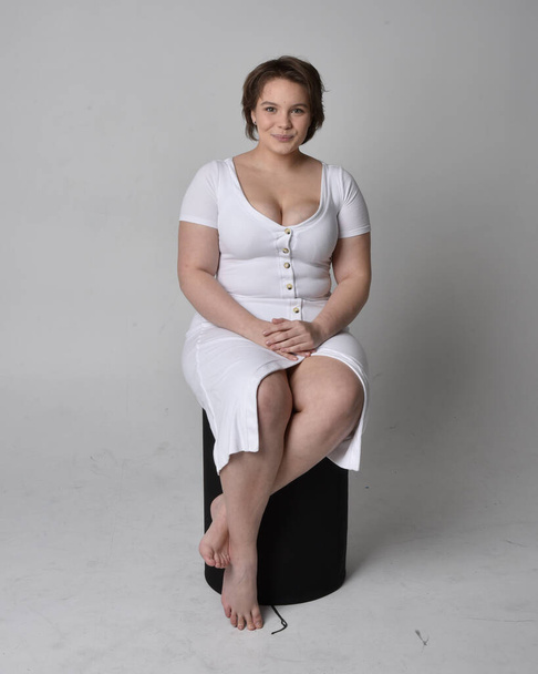 Full length portrait of young plus sized woman with short brunette hair,  wearing tight white body con dress,   sitting pose on chair with gestural hands with light studio background. - Fotoğraf, Görsel