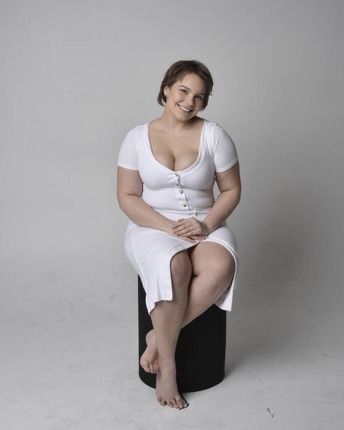 Full length portrait of young plus size woman with short brunette hair, φορώντας στενό λευκό σώμα con φόρεμα, κάθεται θέτουν σε καρέκλα με χειρονομιακή χέρια με φως φόντο στούντιο. - Φωτογραφία, εικόνα