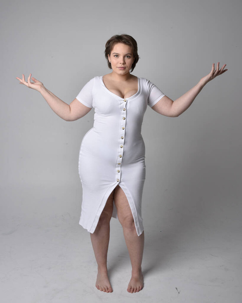 Full length portrait of young plus sized woman with short brunette hair,  wearing a tight white body con dress,   standing walking  posewith light studio background. - Photo, image