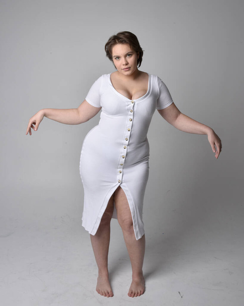 Full length portrait of young plus sized woman with short brunette hair,  wearing a tight white body con dress,   standing walking  posewith light studio background. - Fotó, kép