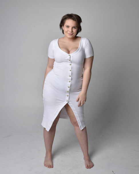 Full length portrait of young plus sized woman with short brunette hair,  wearing a tight white body con dress,   standing walking  posewith light studio background. - Photo, Image