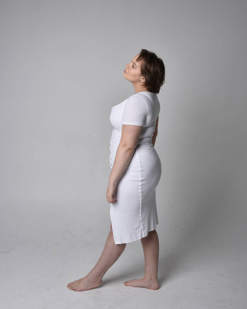 Full length portrait of young plus sized woman with short brunette hair,  wearing a tight white body con dress,   standing walking  posewith light studio background. - Fotografie, Obrázek