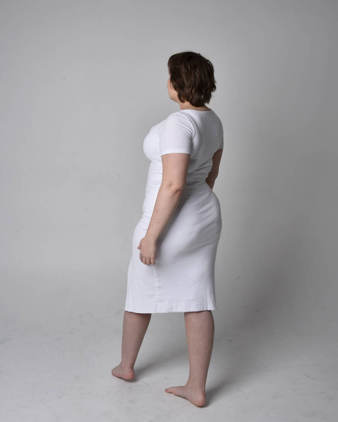 Retrato de larga duración de una joven mujer de tamaño grande con pelo corto morena, con un vestido ajustado cuerpo blanco con, de pie caminando posewith fondo de estudio de luz. - Foto, imagen