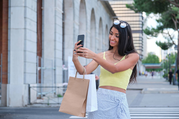 Young latin woman taking a selfie and holding shopping bags at street. - Photo, Image