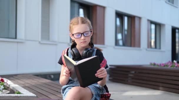  χαριτωμένο καυκάσιο κορίτσι κοκκινομάλλα σε γυαλιά με σακίδιο κάθονται και να διαβάσετε το βιβλίο - Πλάνα, βίντεο