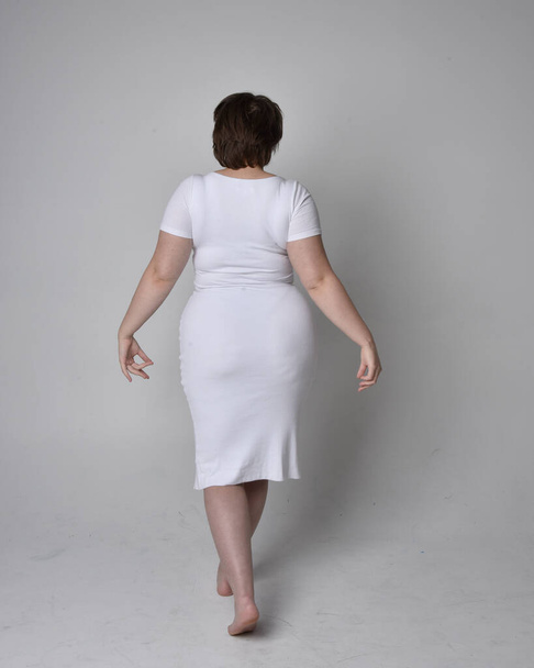 Full length portrait of young plus sized woman with short brunette hair,  wearing a tight white body con dress,   standing walking  posewith light studio background. - 写真・画像