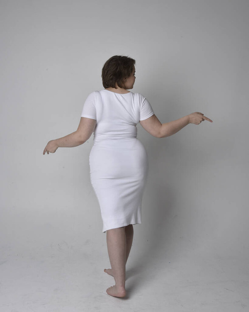 Full length portrait of young plus sized woman with short brunette hair,  wearing a tight white body con dress,   standing walking  posewith light studio background. - Фото, изображение