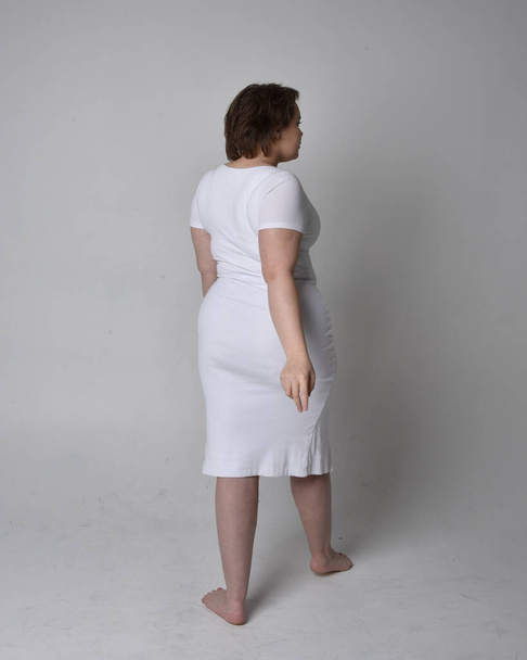 Full length πορτρέτο της νεαρής συν μέγεθος γυναίκα με κοντά καστανά μαλλιά, φορώντας ένα σφιχτό λευκό σώμα con φόρεμα, στέκεται με τα πόδια posewith φως φόντο στούντιο. - Φωτογραφία, εικόνα