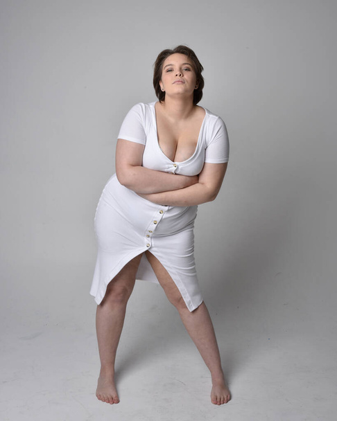 Retrato de larga duración de una joven mujer de tamaño grande con pelo corto morena, con un vestido ajustado cuerpo blanco con, de pie caminando posewith fondo de estudio de luz. - Foto, imagen